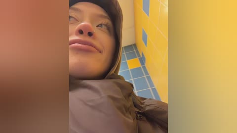 Media: Video of a person with light skin and a nose piercing, wearing a dark hooded jacket, looking upward with a neutral expression. Background features yellow and blue tiled walls.