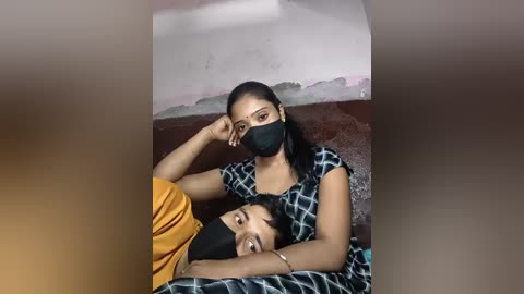 Video of two South Asian women, one in a black mask, lying on a bed with a yellow blanket. The background features a wall with abstract art.