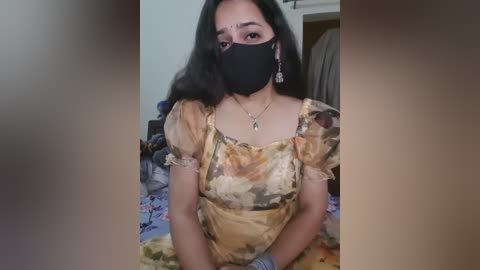 Media: Video of a woman with medium skin tone, wearing a floral dress, black mask, and dangling earrings, seated on a bed with a blue floral sheet.