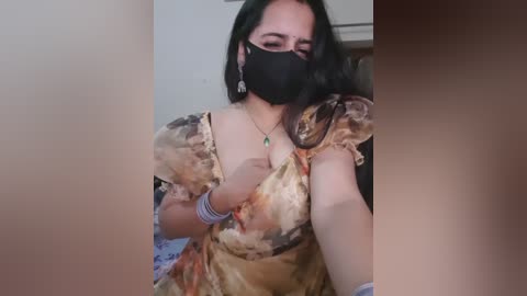 Media: Video of a woman with long black hair, wearing a black mask, floral dress, and bracelets, taking a selfie indoors.
