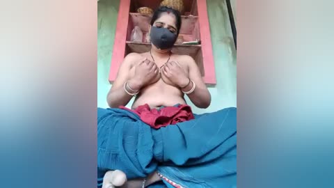 Video of a topless, dark-skinned woman with medium build, wearing a black mask, red skirt, and blue pants, sitting cross-legged, holding her breasts, in a green and pink room with woven baskets.
