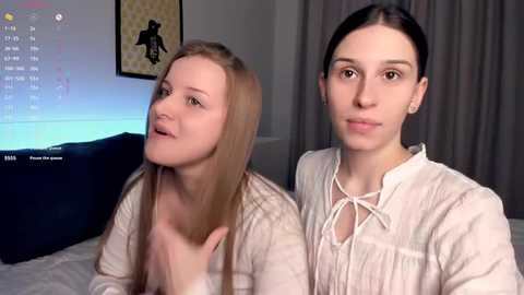 Video of two young women with straight, shoulder-length hair, one blonde and one brunette, sitting on a bed. They wear casual, white tops and appear relaxed, with a TV screen visible in the background.