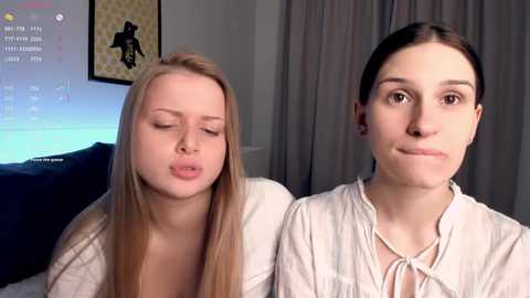Video of two young women with long hair, one blonde, one brunette, sitting close together, smiling, in a cozy room with a TV screen showing a chat window.