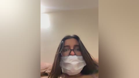 Video of a woman with long dark hair, wearing a white surgical mask and black-framed glasses, lying on a bed with beige walls and a small window emitting light.