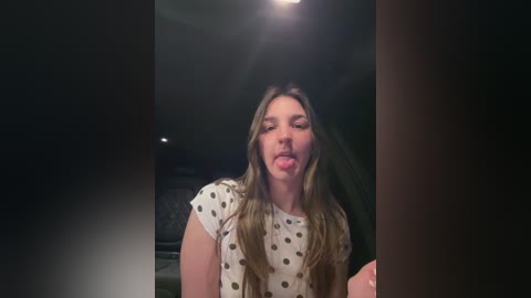 Video of a young woman with long, wavy brown hair, wearing a white polka-dot top, sticking her tongue out playfully in a dimly lit room.