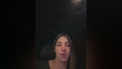A video of a young woman with long brown hair, fair skin, and a neutral expression, seated in a dimly lit car interior.