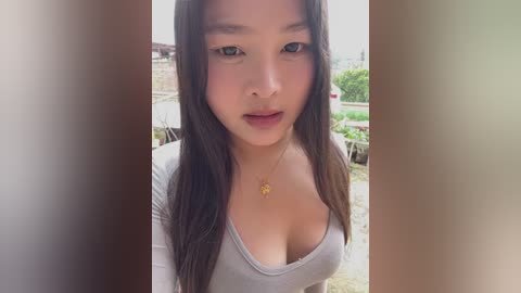 Video of a young Asian woman with long, straight brown hair and fair skin, wearing a low-cut gray top that accentuates her medium-sized breasts. She has a neutral expression, set against a blurred outdoor background with greenery and a wooden fence.