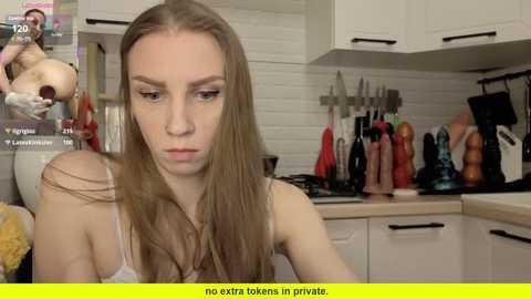 Video of a young woman with long, straight brown hair in a white tank top, sitting in a modern kitchen with colorful utensils on a counter.