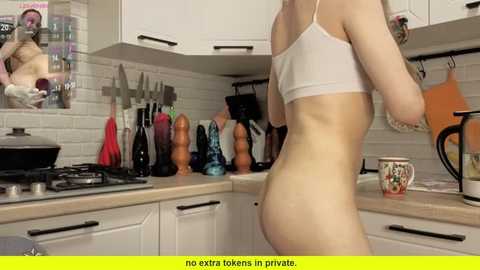 Video of a pale-skinned, slender woman with a white bra and no pants, standing in a modern kitchen with white cabinets, black appliances, and colorful utensils.