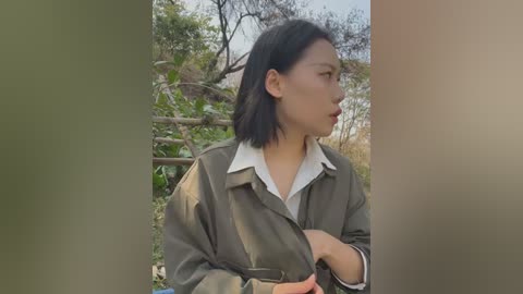 Video of an Asian woman with shoulder-length black hair, wearing an olive green jacket over a white shirt, standing outdoors against a backdrop of trees and a wooden fence.