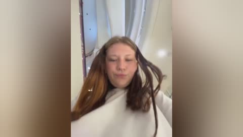 Media: Video of a young woman with light skin and shoulder-length, wavy brown hair, wearing a white salon cape. She stands indoors with a blurred, off-white wall and a window with light filtering through.