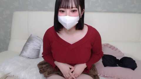 Media: Video of an East Asian woman with straight black hair and bangs, wearing a red V-neck sweater, white mask, and brown skirt, sitting on a white couch with pillows, in a minimalist room.