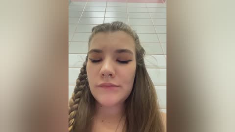Video of a young white woman with light skin, long brown hair styled in two braids, closed eyes, wearing eyeliner, standing in a tiled bathroom with white and pink tiles.