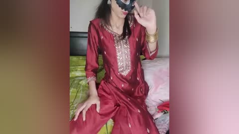 Video of a person wearing a red, gold-embroidered salwar kameez and a black mask, sitting on a bed with pink and yellow bedding in a dimly lit room.