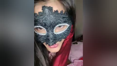 Video of a woman with long dark hair, wearing a black lace masquerade mask with silver embellishments and a red satin cloak. The background is blurred, focusing on her masked face.