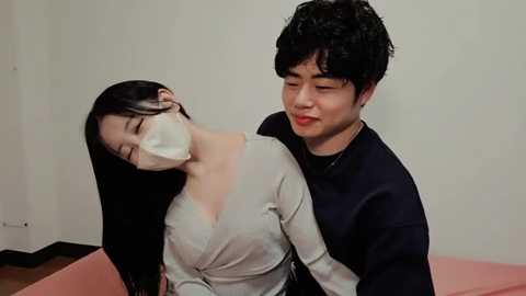 Video of an Asian couple in a medical setting. The man, with curly black hair and a black shirt, gently hugs the woman, who has long black hair and wears a mask, white top, and a pink gown.