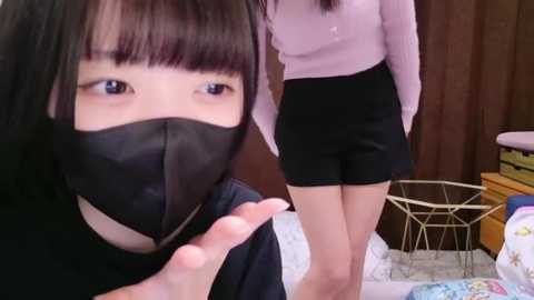 Video of a young Asian woman with black hair and blue eyes, wearing a black face mask, smiling, indoors with wooden walls and a bed in the background.
