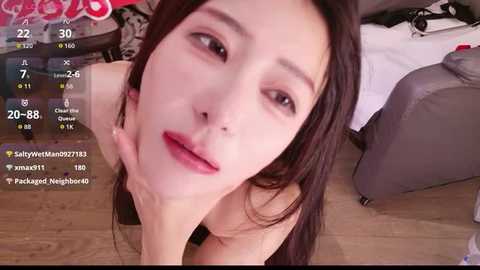 A close-up video of a fair-skinned, attractive woman with long dark hair, light makeup, and red lips, lying on a wooden floor in a bedroom.