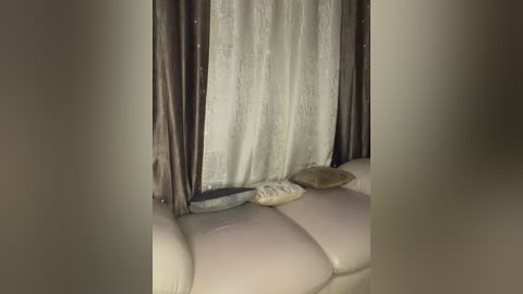 A video of a beige sofa with a light beige pillow and a beige cushion, partially obscured by a sheer white curtain with gold lace trim and matching gold curtains.