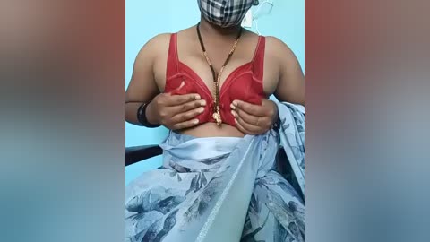 Video of a South Asian woman wearing a red bra, floral sari, and a face mask, seated indoors with light blue walls in the background.