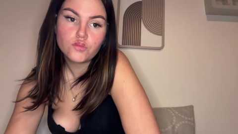 Video of a young woman with fair skin, long dark hair, wearing a black top, puckering her lips, in a minimalist room with beige walls and modern art.