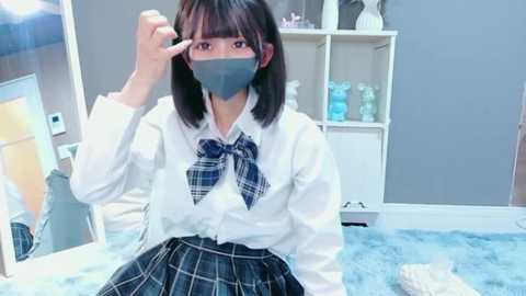 Video of an East Asian girl with straight black hair, wearing a white button-up shirt, blue plaid bowtie, and a grey face mask, adjusting glasses in a modern, brightly lit room with white shelves and blue carpet.