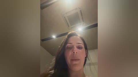 A video captures a close-up of a woman with long brown hair, fair skin, and closed eyes, standing in a dimly lit room with a beige ceiling and an air conditioning unit.