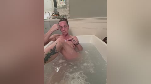 Video of a nude man with light skin, wet hair, and tattoos, sitting in a bathtub filled with water and soap bubbles, in a modern bathroom with white walls and a tiled backsplash.