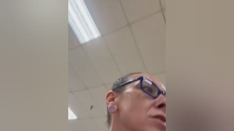 Video of a bald man with glasses and ear gauges in a fluorescent-lit room, looking downward.