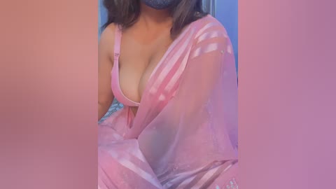 Media: Video of a woman with light brown skin, wearing a pink, sheer, patterned blouse that reveals cleavage, with a blue face mask covering her nose and mouth. Background is blurred, with pink and blue hues.