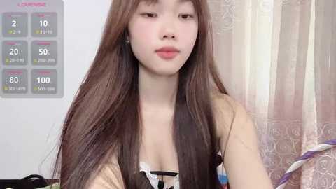 Video of an Asian woman with long, straight brown hair, wearing a beige top, sitting in a room with a whiteboard displaying temperature readings, and a patterned curtain in the background.