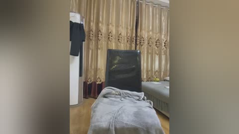 Video of a modern, cluttered living room with beige curtains, a black TV, a gray couch, a white wall, and a folded gray blanket on the floor.