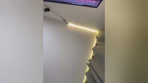 Media: Video of a dimly lit, modern room with a flat-screen TV mounted on the ceiling, emitting a soft yellow glow from beneath a recessed shelf. The walls are plain white, and the room has a minimalist aesthetic.