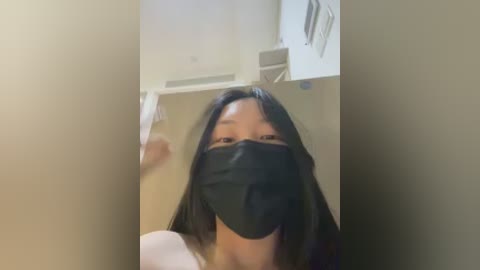 Video of an Asian woman with long black hair, wearing a black face mask, standing in a dimly lit bathroom with beige walls and a white cabinet.