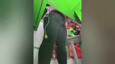 Video of a person wearing tight black leggings and a neon green jacket, bending over in a cluttered room with a green cabinet, a pink jacket hanging, and a gray stool.