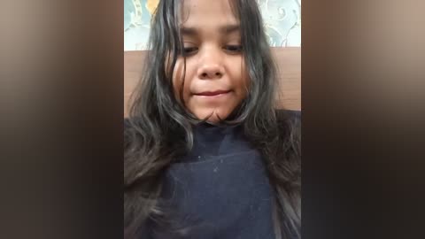 Video of a young Asian girl with wet, dark hair, wearing a black turtleneck, smiling slightly, in a bathroom with a blurred floral wallpaper background.