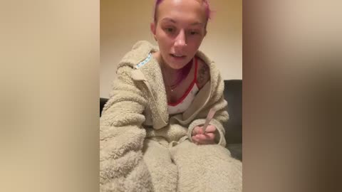 Video of a young woman with pink hair, fair skin, and tattoos, wearing a fluffy beige bathrobe, holding a pink object, in a dimly lit bathroom.