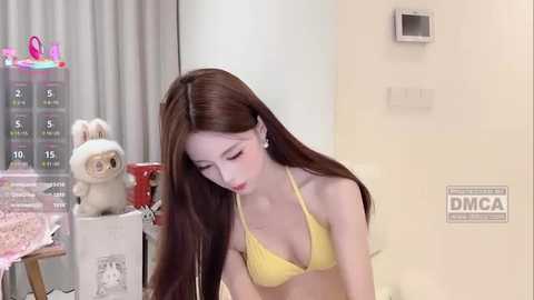 Video of a young Asian woman with long brown hair, wearing a yellow bikini, leaning over a desk with a stuffed animal and a calendar in the background.