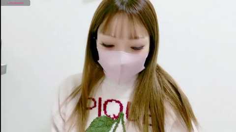Video of an Asian woman with long brown hair, wearing a white sweater with a green leaf design and a pink surgical mask, indoors, looking down.
