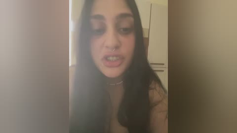 A video of a young woman with long black hair, wearing a choker and a nose ring, smiling with braces, taken indoors in a dimly lit room.