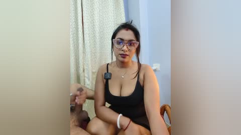 Video of a South Asian woman with medium skin tone, wearing glasses, black tank top, and red bindi, sitting in a chair. Background includes a white curtain and light blue wall.