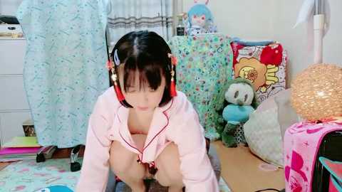 Video of a young Asian woman with short black hair, wearing headphones, in a colorful, cluttered bedroom with plush toys, a pink blanket, and a polka-dotted bedspread.