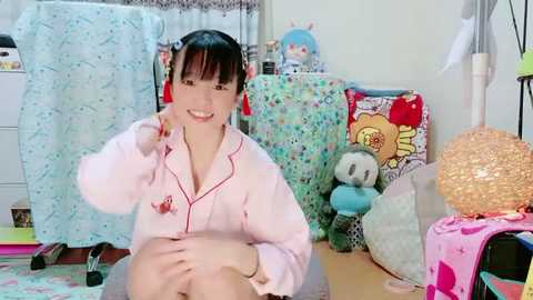 A video of a young East Asian woman with black hair and a pink bunny pajama top, smiling, sitting on a bed with colorful pillows and stuffed animals in a brightly lit, cluttered bedroom.
