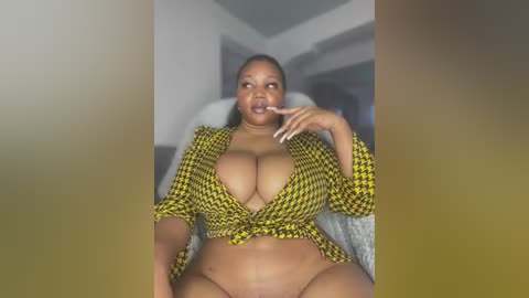 Video of a plus-sized Black woman with large breasts, wearing a yellow-and-black houndstooth top tied at the waist, posing provocatively indoors with a blurred background.