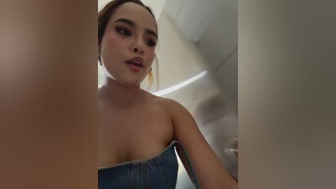 Video of a young woman with light skin, brown hair, and full lips, wearing a strapless denim top, in a dimly lit, modern bathroom with white tiles.