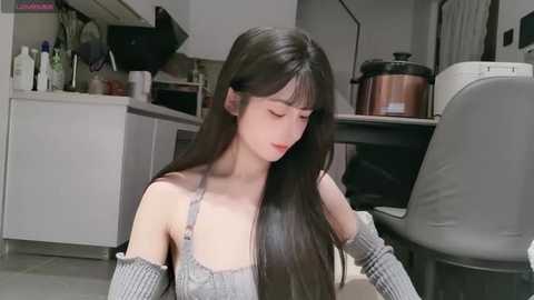 Media: Video of a young Asian woman with long, straight black hair, wearing a grey off-shoulder sweater, seated in a modern kitchen with white cabinets, a copper pot, and grey leather chair.