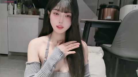 Video of an East Asian woman with long black hair, pale skin, and a slender build, wearing a gray off-shoulder sweater, sitting on a white couch in a modern kitchen.