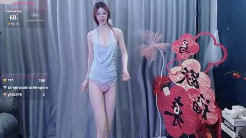 Video of a slender, fair-skinned woman with long brown hair, wearing a revealing white halter top and pink lace panties, standing in front of a heart-shaped, red-pink floral arrangement.