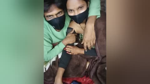 A video captures two women, one with light skin, wearing a green shirt, and the other with dark skin, wearing a black shirt, both with black masks, hugging tightly in a dimly lit room.