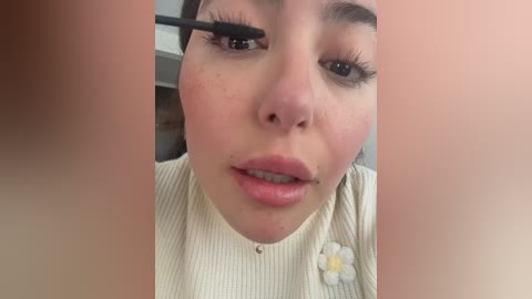 A close-up video of a young woman with fair skin, large eyes, and light pink lips, applying black mascara to her left eye, wearing a cream-colored top with a daisy pin.